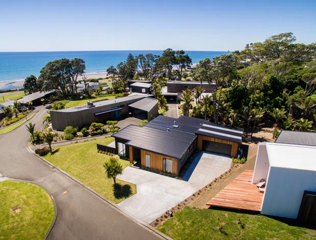 Aerial view of a contemporary coastal residential development beside a sandy beach and ocean. Several low-profile homes feature dark metal roofs, dark cladding, timber accents, large glazing, landscaped gardens, driveways, and decks.