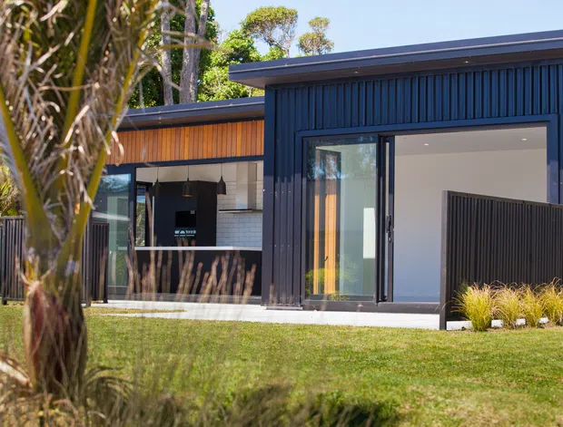 Exterior view of a contemporary single-storey home with dark vertical cladding, broad glazed sliding doors, and a partially visible outdoor kitchen or entertaining area. Warm timber cladding accents the recessed section, while landscaped lawn and tropical planting frame the facade.