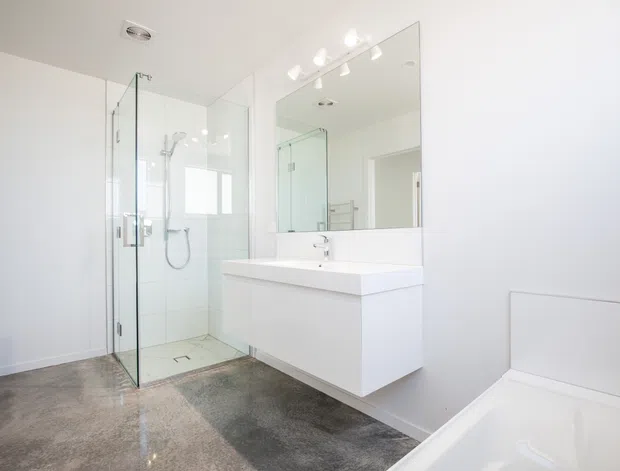 Bright contemporary bathroom with a frameless glass walk-in shower, wall-mounted white vanity with integrated basin, large mirrored cabinet, freestanding bathtub edge, and polished grey stone-look flooring. The room has white walls, recessed fittings, and a clean minimalist appearance.