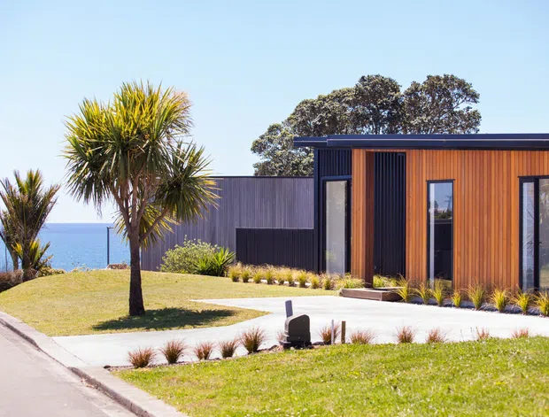 A contemporary single-storey coastal house viewed from the street, with a flat roof, dark vertical cladding, warm timber-look facade panels, large black-framed windows, and a landscaped front lawn. The ocean is visible beyond the property, with palm-like plants and low ornamental planting around the house.
