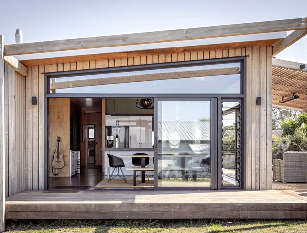 Exterior view of a compact timber-clad pavilion or extension with a raised timber deck, large sliding glass doors, exposed structural posts and beams, and a pergola-covered outdoor sitting area. Through the glazing, a contemporary kitchen and dining area are visible. The design uses natural weathered timber and strong indoor–outdoor connection.