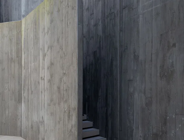 A minimalist architectural stairway enclosed by tall dark grey concrete walls. A lighter concrete partition forms a sculptural angled edge, concealing the stair flight. Large pale floor tiles contrast with the heavily textured vertical concrete surfaces, creating a stark, dramatic composition with strong natural light and shadow.