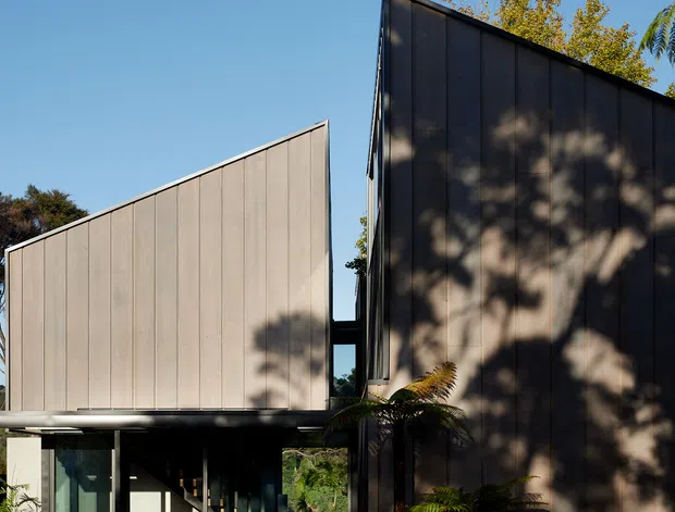 Exterior view of a contemporary split-level residence with two sharply angled, wing-like volumes clad in dark and light vertical panels. A glazed entrance connection sits between the forms, with visible interior stairs and timber furnishings. Tropical and subtropical planting frames the paved approach, while strong sunlight casts tree shadows across the facade.