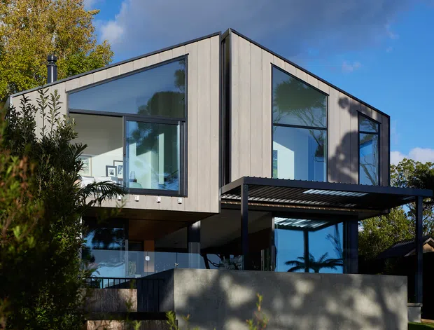 Exterior view of a contemporary multi-level house with angular rooflines, vertical grey cladding, extensive floor-to-ceiling glazing, glass balustrades, and a dark metal pergola or carport-like canopy. The home is set within landscaped suburban gardens under a partly cloudy sky.