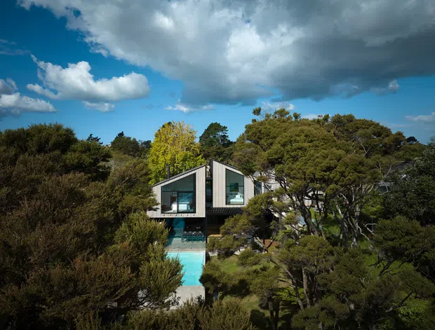A modern elevated house partially concealed within dense native vegetation. The facade features angular, asymmetrical roof forms, dark cladding, large glazed openings, and a turquoise swimming pool extending in front of the building. Dramatic clouds and mature trees surround the secluded property.