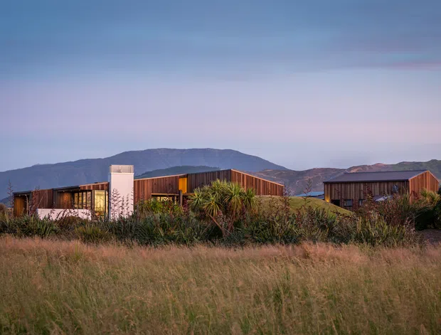 A low-rise contemporary residential complex viewed from a grassy landscape at dusk. The buildings feature long horizontal forms, dark timber-toned cladding, extensive glazing, and a prominent angled roofline, set against a mountainous backdrop and landscaped native planting.