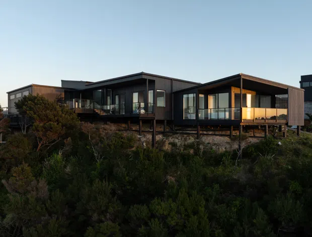 A modern multi-wing residential house elevated on slender supports above a dense natural coastal-looking landscape. The dark-clad building has a mix of low-pitched and flat roof forms, extensive floor-to-ceiling glazing, timber-toned wall sections, balconies, and warm interior lighting visible at dusk. The image is an exterior architectural view with the surrounding vegetation in the foreground.