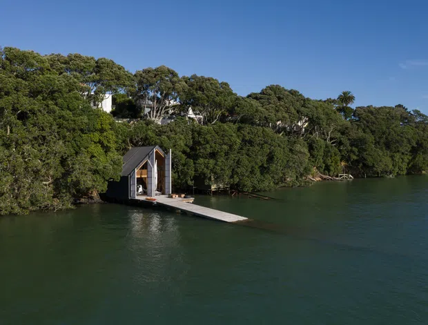 Aerial exterior view of a small dark-clad, steeply gabled structure positioned at the edge of calm water, connected to shore by a narrow floating or projecting walkway. Dense mature trees surround the waterfront site, with glimpses of a larger residence among the vegetation. Clear blue sky and tranquil water create a secluded coastal or lakeside setting.