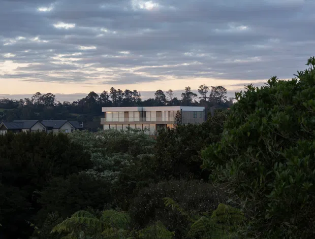 Dusk exterior view across dense garden vegetation toward a modern, low-rise residential building with a long rectangular form, pale facade, extensive horizontal glazing, and a flat roof. Dark pitched-roof homes are visible in the background, beneath a cloudy evening sky.