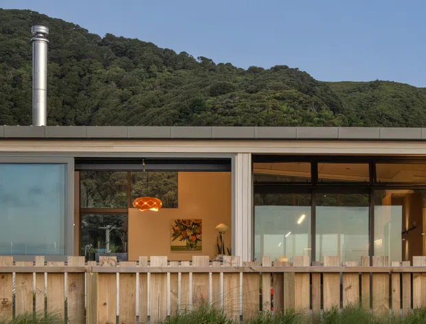 A low-rise modern house viewed from outside, set against a densely wooded hillside. The facade has extensive floor-to-ceiling glazing, pale vertical structural elements, a flat roof with a metal chimney, and a timber slat deck or balustrade in the foreground. Warm interior lighting and furnishings are visible through the windows, with natural grasses and planting around the home.