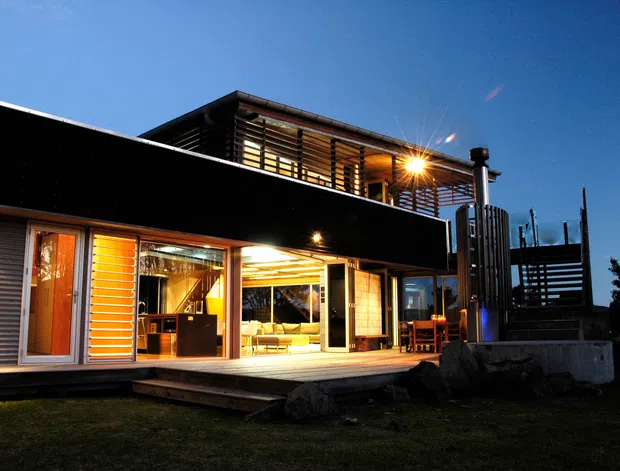 Twilight exterior view of a contemporary two-storey residence with dark horizontal cladding, extensive timber-framed glazing, illuminated open-plan living areas, a projecting upper-level balcony or deck enclosed by horizontal timber screening, and an exterior stair or platform. The home sits in a dark landscaped setting beneath a clear blue sky.