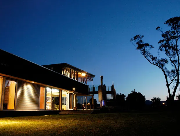 A contemporary single-storey house photographed at dusk, with a long low roofline, extensive illuminated glazing, timber or metal horizontal screening, and a raised upper pavilion or deck element. The building sits in a dark landscaped setting with silhouetted trees and a broad lawn. Warm interior and exterior lighting contrasts with the deep blue evening sky.