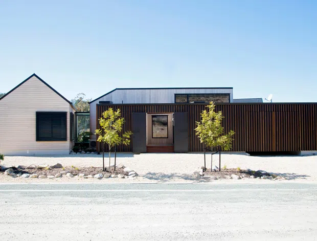 A single-storey contemporary residential building viewed from the street, composed of a small gabled volume on the left and a larger flat-roofed structure behind a dark vertical-slatted facade. The exterior uses pale horizontal cladding, dark timber-toned screening, and simple rectangular glazing and entry elements. A gravel forecourt with young trees and low rock edging sits in front of the house.
