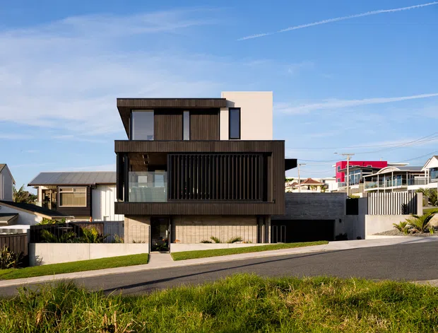 Exterior street view of a contemporary multi-storey residence with a strongly rectilinear composition. The facade combines dark vertical timber-like cladding and screening, pale rendered wall surfaces, extensive glazing, and a low-profile flat roof. A recessed ground-level entry and covered outdoor area are visible, with neighbouring suburban homes surrounding the property.