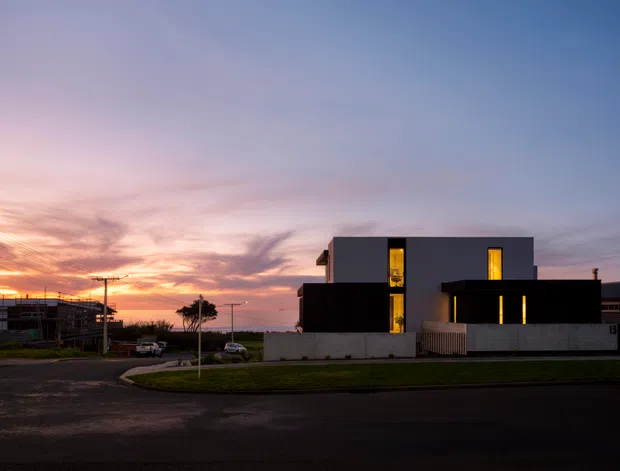 A modern two-storey dark-clad house photographed at dusk from across a suburban street. The building has a strong rectilinear composition, flat rooflines, narrow vertical illuminated windows, and a low pale boundary wall. A neighbouring structure under construction is visible to the left, with a sunset sky behind the homes.