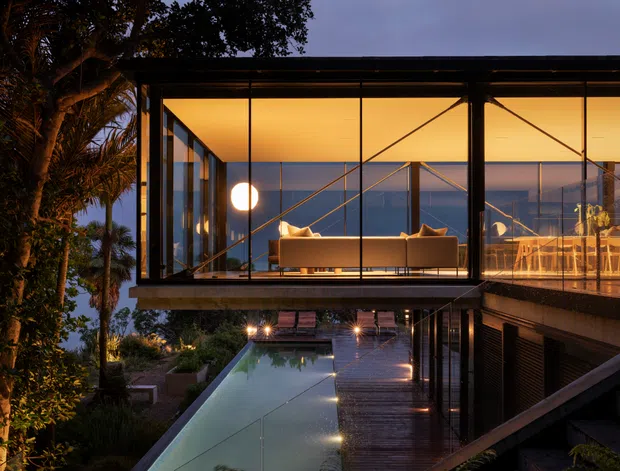 Twilight exterior view of a modern elevated glass pavilion beside a long narrow pool. The transparent upper level reveals a softly lit living room and dining area, while dark structural framing, extensive glazing, timber decking, and landscaped planting create a refined indoor-outdoor composition.