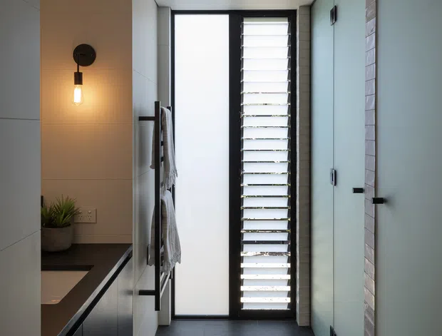 A narrow, contemporary bathroom with a dark tiled floor, pale large-format wall tiles, dark built-in vanity cabinetry and a black benchtop. A frameless glass shower enclosure occupies the right side, while a tall frosted window and black louvred window provide filtered natural light. A wall-mounted black pendant light, towel rail, and small potted plant add detail.