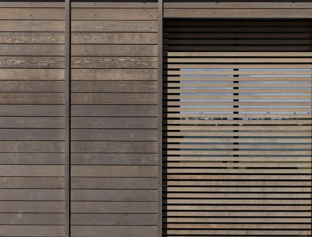Close-up exterior view of a contemporary timber-clad facade with horizontal weathered wood boards, dark vertical framing, a corrugated metal roof, and a large horizontal slatted screen opening. Pale concrete paving is visible at the base, with limited views through the screen to the exterior beyond.