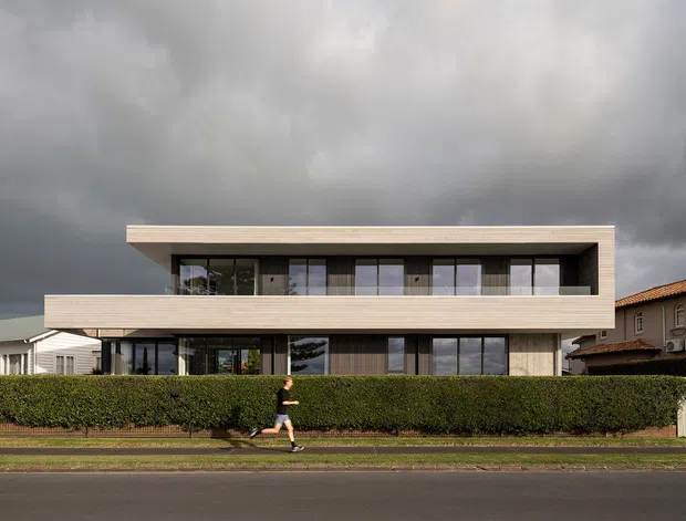 Street-facing view of a contemporary two-storey residence with a long, flat roof and strongly horizontal composition. The facade combines pale horizontal cladding, extensive dark-framed glazing, recessed upper-level balconies or covered terraces, and a neatly clipped hedge along the front boundary. Neighbouring homes and a dramatic overcast sky are visible in the background.