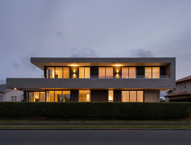 Twilight street-facing view of a large contemporary two-storey residence with a strongly horizontal composition, flat roof, dark timber-like cladding, extensive warm-lit glazing, projecting upper-level forms, and a neatly trimmed hedge along the frontage.