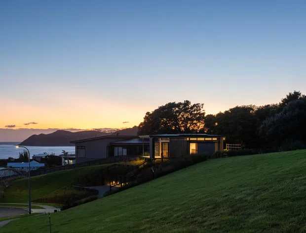 A low-profile modern house sits on a sloping grassy site overlooking the sea at sunset. The single-storey building has dark horizontal cladding, extensive glazed openings, and warm interior lighting, with nearby residential structures and a coastal headland visible in the background.