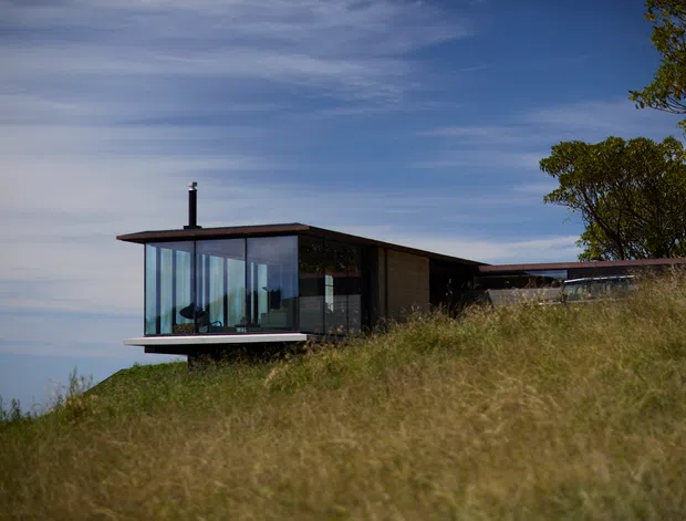 A low, contemporary house sits on a grassy elevated site, with a broad flat roof, dark structural framing and extensive floor-to-ceiling glazing at the projecting living area. A chimney rises from the roof, while the surrounding dry grasses and trees partially screen the building.