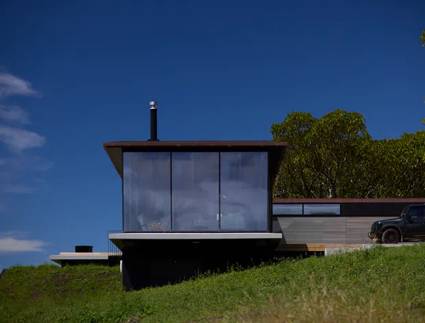 A modern elevated house set on a grassy sloping site, photographed from the exterior under a deep blue sky. The compact upper volume has extensive floor-to-ceiling glazing, dark structural elements, and a thin projecting roof with a black flue. A low horizontal wing extends behind it with muted timber or composite cladding and narrow strip windows; a vehicle is parked beside the building. The composition emphasizes the cantilevered, glass-fronted form and connection to the landscape.