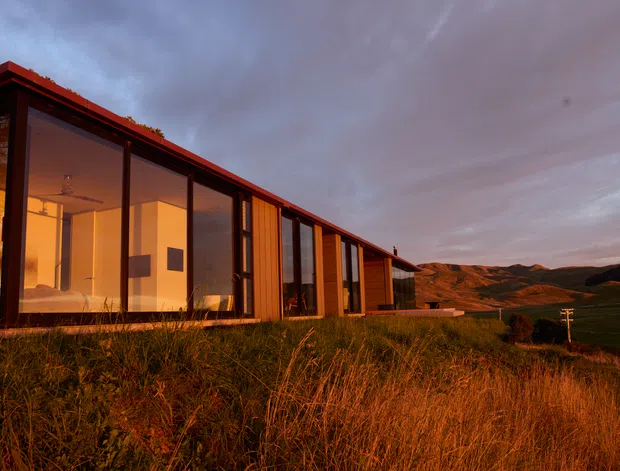 A low, elongated modern dwelling set on a grassy, elevated rural site at dusk. The building features extensive floor-to-ceiling glazing, dark slender window frames, warm timber-toned vertical cladding, and a shallow pitched or low-slope roof. Warm interior lighting contrasts with the cool evening sky, while rolling hills are visible beyond the house.