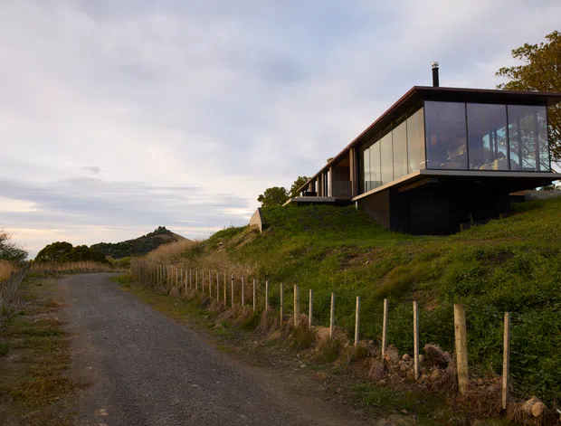 A contemporary dark-clad house sits elevated on a grassy coastal or rural slope beside a gravel access track. The building has a dramatic projecting glazed living volume, extensive floor-to-ceiling windows, a low pitched roof, and timber-toned structural framing. The surrounding landscape includes fencing, dune-like vegetation, and distant low hills under an overcast sky.