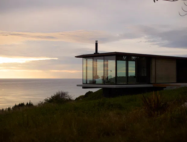 A modern coastal house perched above the ocean, photographed at sunset. The single-storey structure has a flat roof, extensive floor-to-ceiling glazing, dark slim window frames, and a projecting glazed living space overlooking the water. Native grasses and low coastal vegetation surround the building.