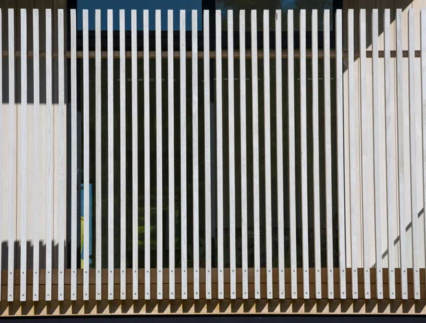 Close-up architectural view of a contemporary vertical slatted screen or fence. Repeated slender white and dark timber-toned members create a strong linear pattern, with a partially visible light-coloured structure behind and greenery seen through the gaps. The image focuses on the facade or screening element rather than an identifiable room.