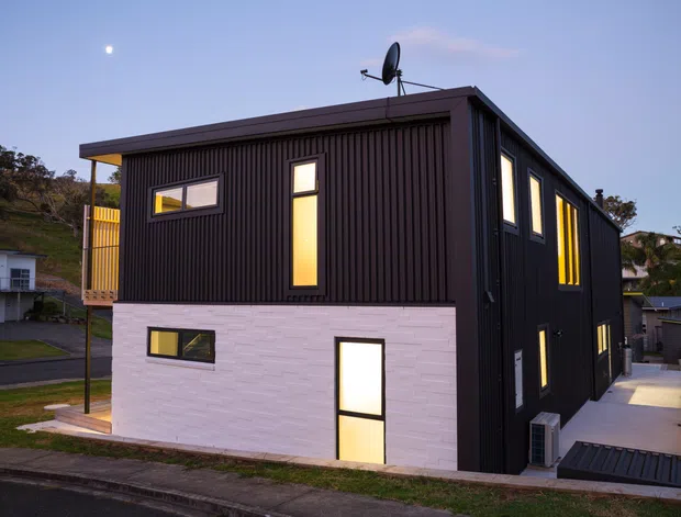 Exterior view of a compact two-storey contemporary dwelling with dark vertical cladding, a pale masonry lower level, and multiple narrow illuminated windows. The building has a simple flat or very low-pitched roof, a projecting upper volume, and a side access path with steps and a small external air-conditioning unit. A neighbouring residential streetscape and a small balcony are visible.