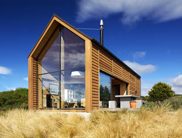 Exterior view of a compact contemporary timber-clad house set within dry ornamental grasses. The building has a steep gabled roof, extensive floor-to-roof glazing on the front elevation, horizontal timber slat cladding, a projecting covered outdoor area, and a visible black flue. The rural landscape and strong daylight emphasize a simple cabin-like architectural form.