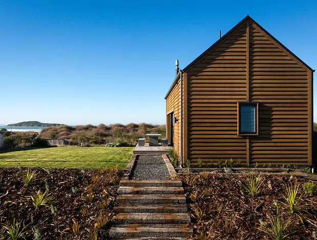 Exterior view of a compact single-storey gable-roofed building clad in horizontal timber boards. A gravel path and timber steps lead through landscaped planting to the entrance, with open coastal scenery visible beyond.