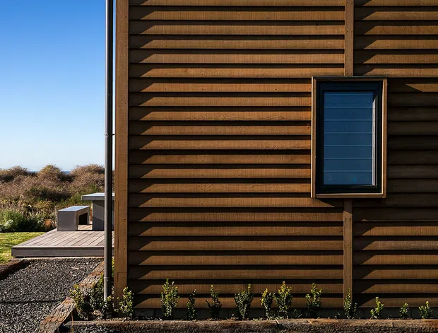 Close-up exterior view of a contemporary building clad in dark-stained horizontal timber boards with a projecting, louvred-like profile. A small dark-framed window punctuates the facade, while a timber deck, gravel path, low planting, and open coastal-looking landscape are visible beside the structure.