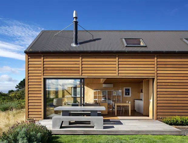 Exterior view of a contemporary timber-clad dwelling with a dark corrugated pitched roof, roof skylights, and a black flue. Large glazed sliding doors open onto a timber deck with a built-in concrete picnic-style table and seating. The open interior reveals a compact kitchen and dining area, while landscaped grasses and shrubs surround the house.