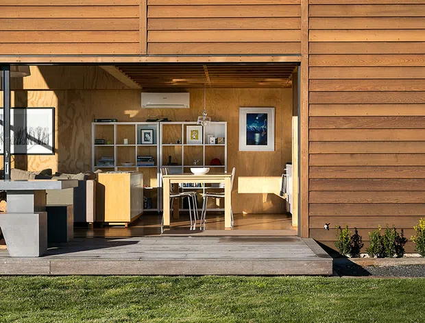 Exterior view of a compact timber-clad building with a large open glazed opening revealing a combined living and dining area. The interior features warm plywood walls, open shelving, a small dining table, and contemporary furnishings, opening onto a timber deck and lawn.