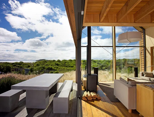 A contemporary indoor-outdoor living and dining area opening onto a deck, with full-height glazing framing a lush coastal landscape. The interior has a warm timber-lined ceiling and floor, exposed structural framing, a neutral lounge suite, and a built-in timber cabinet. Outside, a long concrete dining table with matching bench seating occupies the deck.