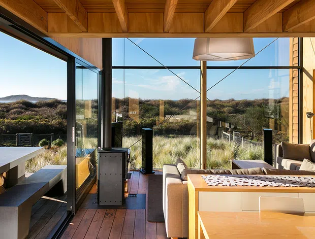 A bright contemporary living and dining space opening onto a coastal landscape through extensive floor-to-ceiling glazing. The room features warm timber ceiling beams and wall panelling, timber flooring, a built-in dining bench and table, upholstered seating, and a sculptural pendant light. Native dune vegetation and the ocean are visible beyond the glazing.