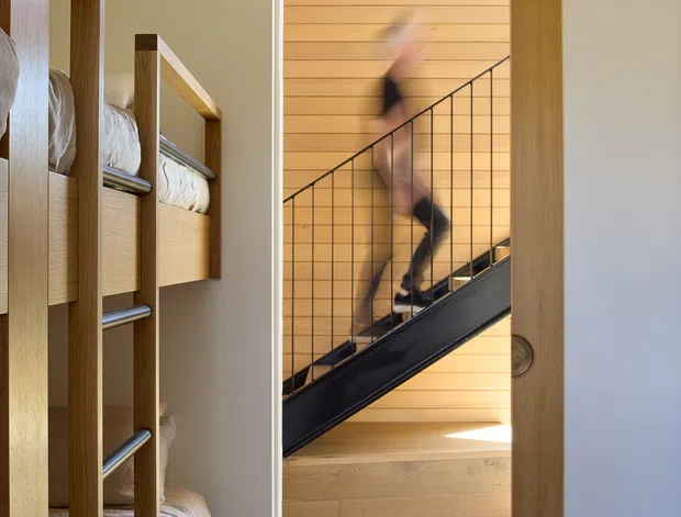 A compact bunk bedroom viewed toward an open doorway leading to a stairwell. Built-in timber bunk beds with white bedding line the left side, while the adjoining stair features warm horizontal timber wall cladding, a black metal balustrade, and timber flooring. The room has soft neutral walls, carpet flooring, and a simple contemporary fit-out. A blurred person is moving on the stairs.