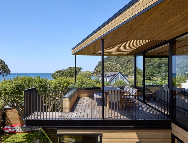 Exterior architectural photograph of a contemporary multi-level coastal home with extensive timber cladding and soffits, dark slender metal framing, and large glazed openings. The image focuses on an elevated outdoor terrace with black metal balustrades, dining furniture, and a slatted timber pergola/ceiling. Ocean, landscaped gardens, and neighbouring rooflines are visible beyond the deck.