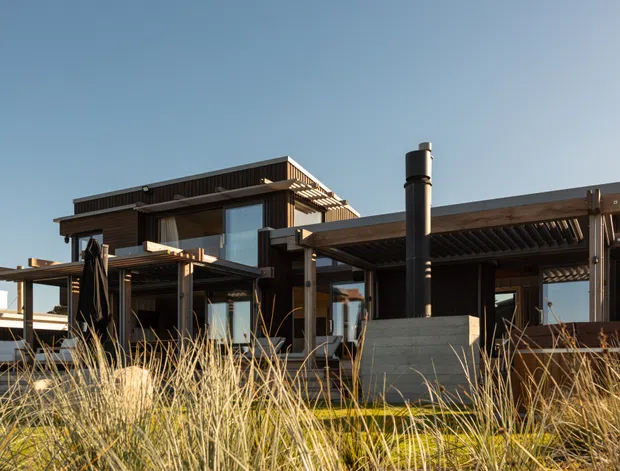 Exterior view of a contemporary multi-level home with strong horizontal roof planes, dark timber or metal cladding, extensive glazing, pergola-like outdoor structures, and a prominent cylindrical chimney. Native ornamental grasses occupy the foreground, and the building is shown under a clear sky.
