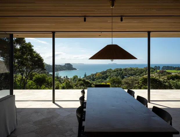 A contemporary dining area opening onto a broad terrace, with a long dark dining table, black chairs, and a large sculptural pendant light beneath a timber-lined ceiling. Full-height glazed sliding doors frame expansive coastal and ocean views, while the pale irregular stone terrace flooring continues the indoor-outdoor connection.