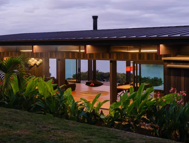 Exterior dusk view of a low-slung modern home with a broad dark pitched metal roof, timber-clad fascia and exposed timber structure. Extensive floor-to-ceiling glazing opens onto a warm timber-floored living area with a central suspended fireplace, while lush tropical planting fills the foreground. The image has a dramatic, warm evening atmosphere and suggests a garden-facing or coastal setting.
