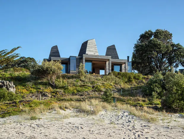 A low, contemporary coastal house viewed from a sandy beach. The dark timber-clad building sits on a gently vegetated dune, with strong geometric roof forms, large glazed openings, and sheltered outdoor terraces. Native coastal planting and rocky landscaping surround the structure under a clear blue sky.