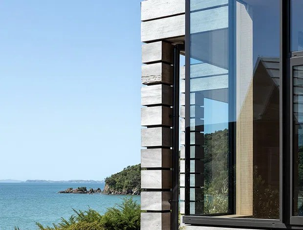 Close exterior view of a contemporary coastal house beside a blue waterfront. The facade combines broad horizontal weathered timber cladding with dark framed floor-to-ceiling glazing and projecting timber elements. A rocky, vegetated shoreline and clear sky are visible beyond the building.