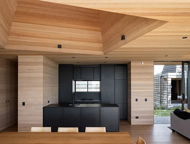 A refined open-plan kitchen and living area featuring extensive light timber wall, ceiling, and built-in cabinetry finishes. The kitchen has a matte black island and dark cabinetry, integrated appliances, and a compact horizontal window. A sculptural recessed timber ceiling detail adds architectural interest, while full-height glazing opens toward an outdoor area and brings in natural light. The foreground includes a timber dining table, and part of a grey sofa is visible to the right.