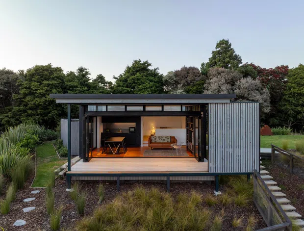 Exterior view of a compact contemporary pavilion-like dwelling set within landscaped grounds. The building has dark vertical cladding, a low flat roof, extensive glazed sliding openings, and a raised deck with an open living area visible inside. Native grasses and dense planting surround the structure, with a timber walkway leading to it.