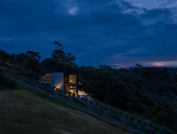 A small modern house is illuminated at dusk on a steep, heavily vegetated hillside. The compact structure has dark vertical cladding or walls, large warm-lit windows, and a simple contemporary form, with a long railing or access path descending across the slope. Dense forest fills the surrounding landscape beneath a dramatic blue evening sky.