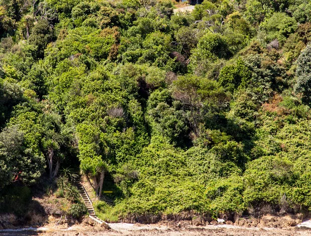 Exterior view of a modern dark-clad house positioned high on a densely vegetated coastal or waterfront hillside. The building has a simple rectangular form, large windows, vertical ribbed dark metal cladding, and timber-toned lower wall surfaces. A steep stairway descends through the vegetation toward a sandy shoreline and calm water, with a kayak in the foreground.